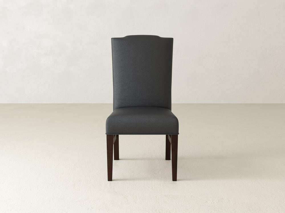 A tall, dark grey upholstered chair with a high back and wooden legs stands on a light beige floor against a textured, off-white wall. The chairs design is simple and elegant.