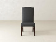 A tall, dark grey upholstered chair with a high back and wooden legs stands on a light beige floor against a textured, off-white wall. The chairs design is simple and elegant.