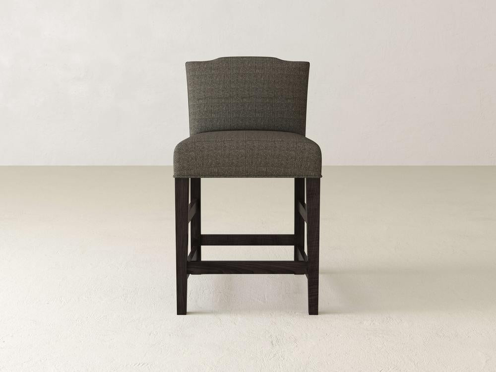 A Hadley Upholstered Stool by James & James, featuring dark wooden legs, stands on a light floor against a plain, light-textured backdrop.