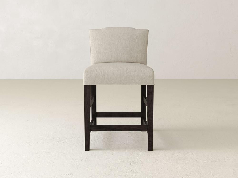 The Hadley Upholstered Stool by James & James, featuring beige fabric and dark wooden legs, exudes sophistication on a light floor against a white wall, enhancing dining experiences with its elegant design.