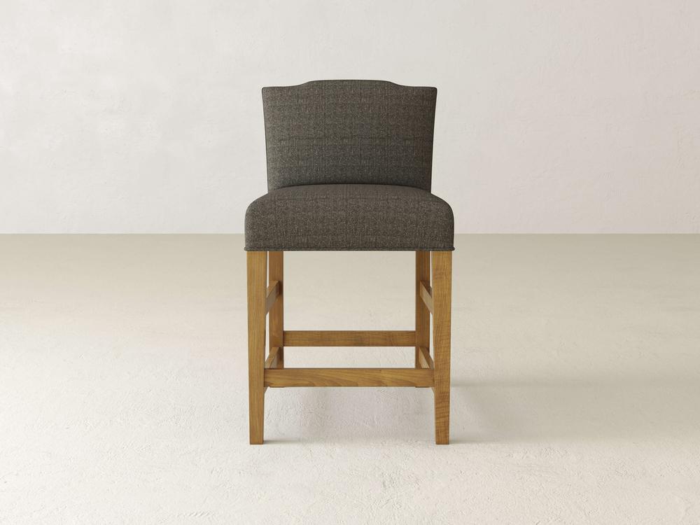 The Hadley Upholstered Stool by James & James, with its plush dark gray fabric seat and backrest, stands elegantly on a light beige floor against a plain wall, enhancing any dining experience with refined simplicity.