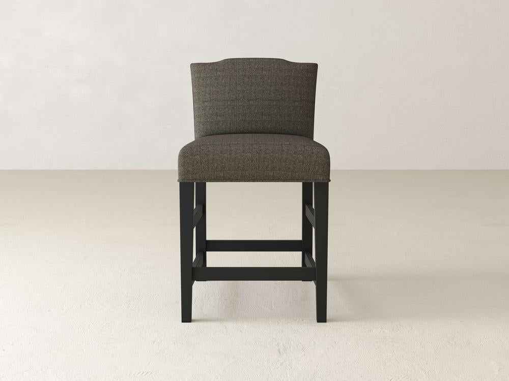 The Hadley Upholstered Stool by James & James features a modern design with an upholstered fabric cushion and black wooden legs, resting on a light beige floor against an off-white backdrop. Its subtly curved high backrest adds comfort and style to any dining experience.
