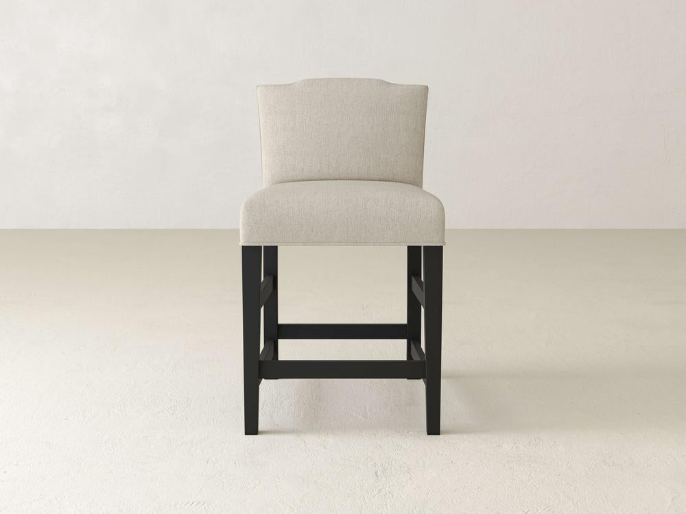 The Hadley Upholstered Stool by James & James features a beige design and cushioned backrest. It stands on dark wooden legs, centered on a light floor with a soft, light-colored background.