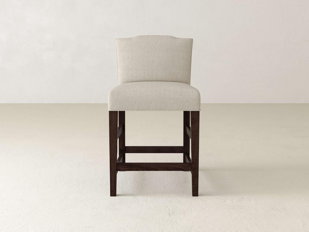 The Hadley Upholstered Stool by James & James features a beige fabric and dark wooden legs, set against a plain light floor and pale wall.