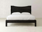 The Harlow Bed - Charred Ember on Alder by James+James features a curved headboard and black wooden frame, shown here with white bedding and pillows in a minimal, modern room.