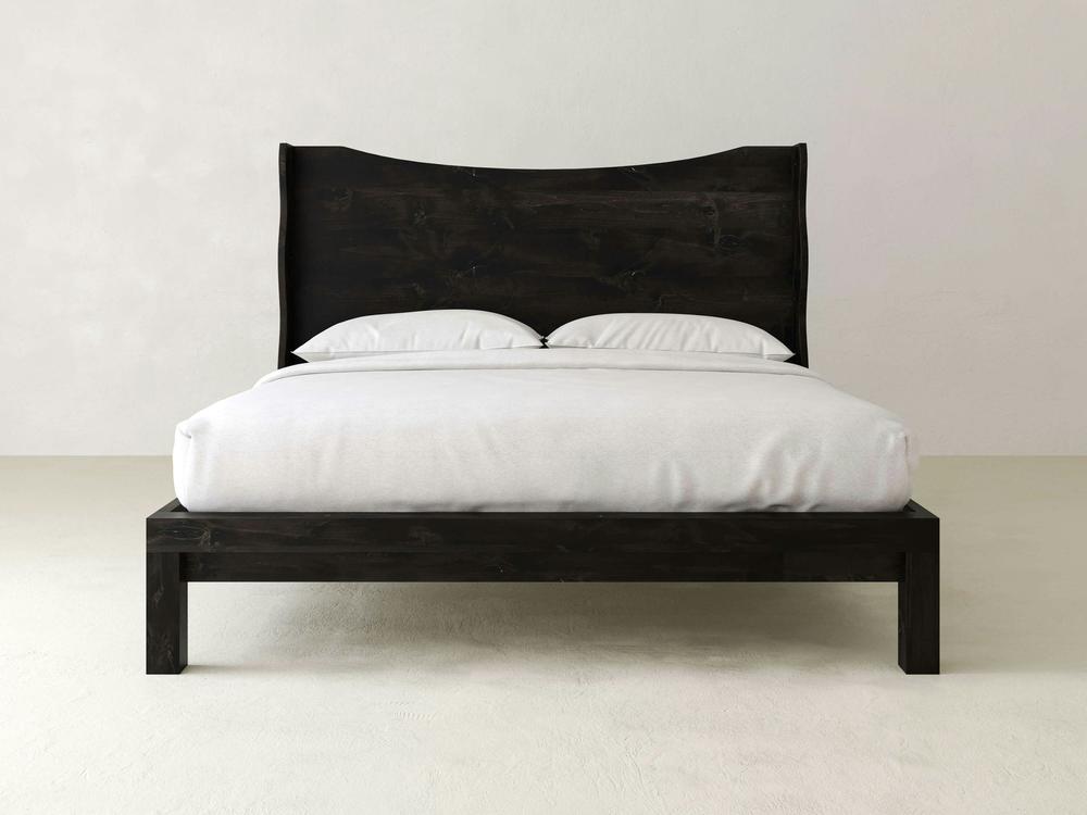 The Harlow Bed - Charred Ember on Alder by James+James features a curved headboard and black wooden frame, shown here with white bedding and pillows in a minimal, modern room.