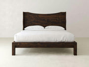 The Harlow Bed - Tobacco on Alder by James+James features a high headboard and dark, rustic finish, styled with white bedding and two white pillows against a plain, light-colored wall.