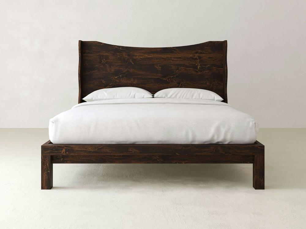 The Harlow Bed - Tobacco on Alder by James+James features a high headboard and dark, rustic finish, styled with white bedding and two white pillows against a plain, light-colored wall.