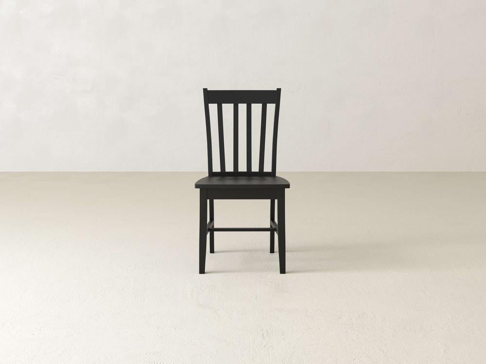 A wooden chair with vertical slats on the backrest sits on a light-colored, smooth floor against a plain, light wall. The design is simple and minimalist.