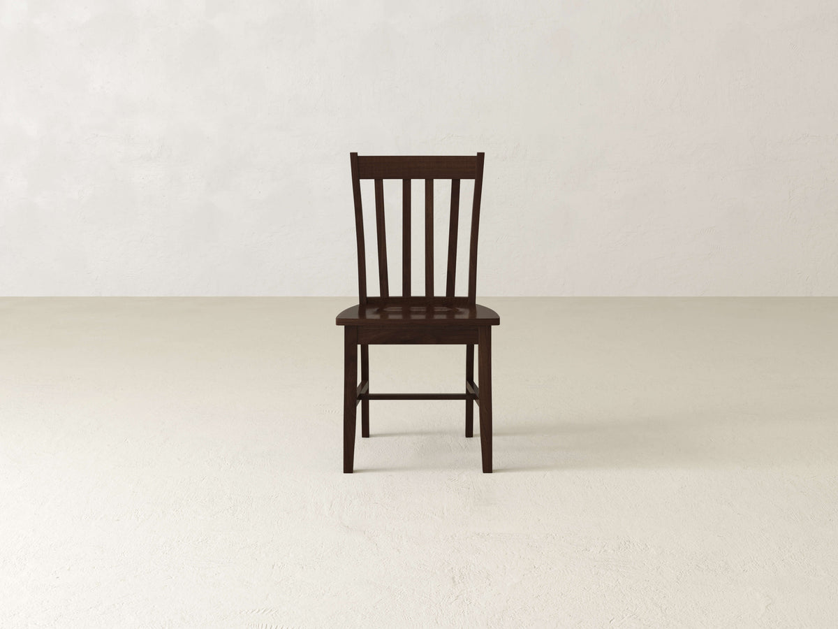 The Henry Dining Chair - Tobacco by James+James features a wooden design with a high backrest adorned with vertical slats. The chair boasts a dark wood finish and four straight legs, showcasing simple and sturdy construction. Its flat seat complements the classic and minimalist design, as shown from an angled rear view.