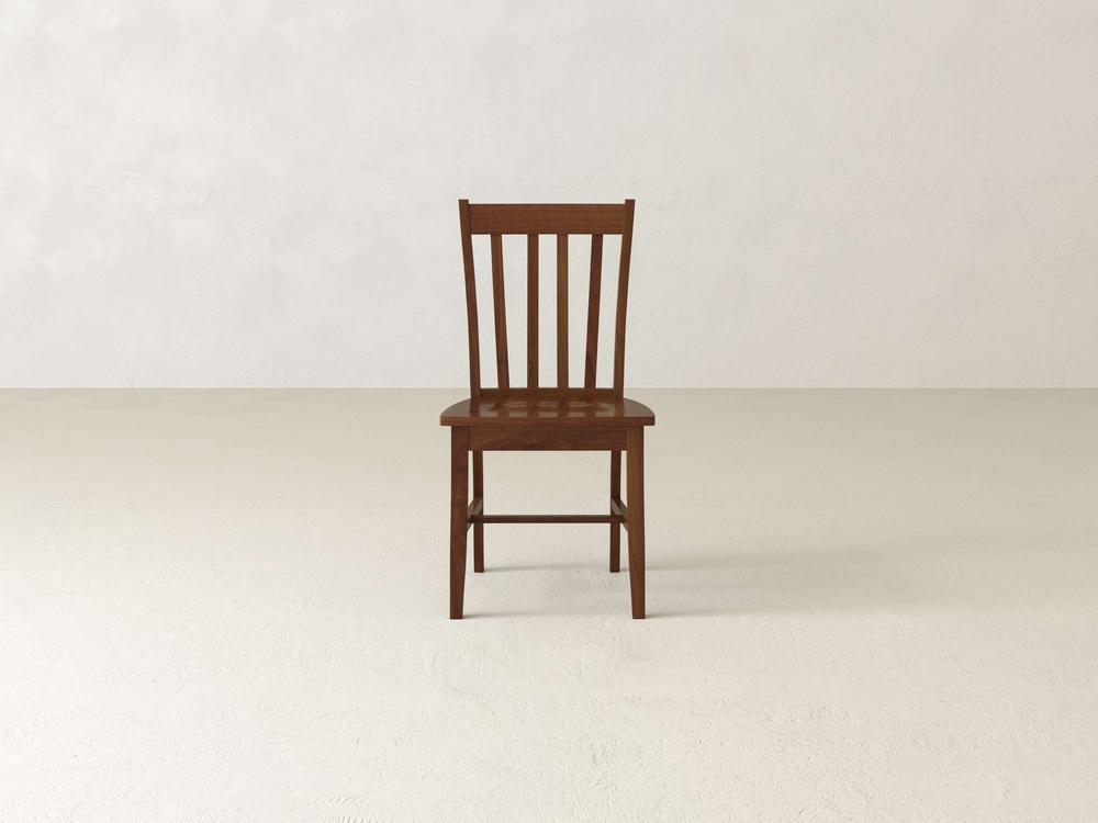 Wooden chair with a simple design and vertical slats on the backrest, placed in the center of an empty room with a plain beige floor and wall.