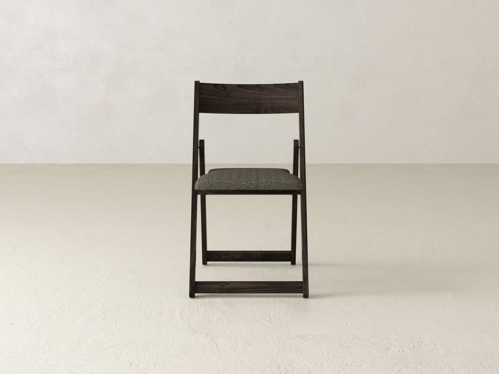 A simple wooden folding chair with a black frame and a gray cushioned seat, placed in an otherwise empty room with a beige floor and off-white walls. The chair faces forward and is centered in the image.