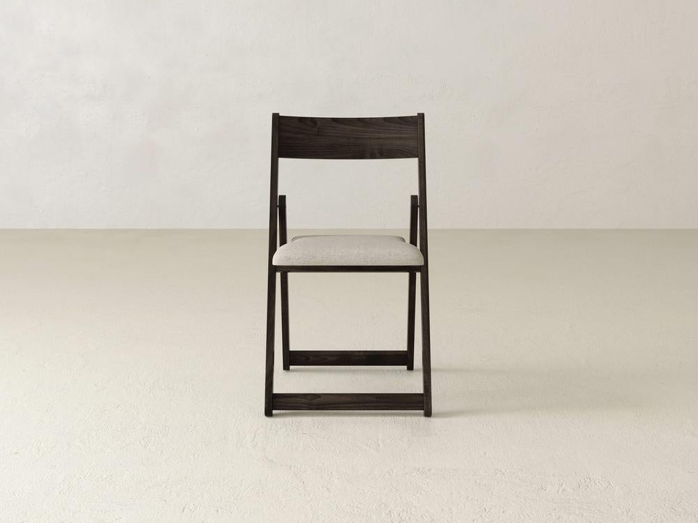 A wooden folding chair with a light gray cushioned seat is placed against a plain, light-colored background. The chair is viewed from the front, showing its simple and minimalist design.