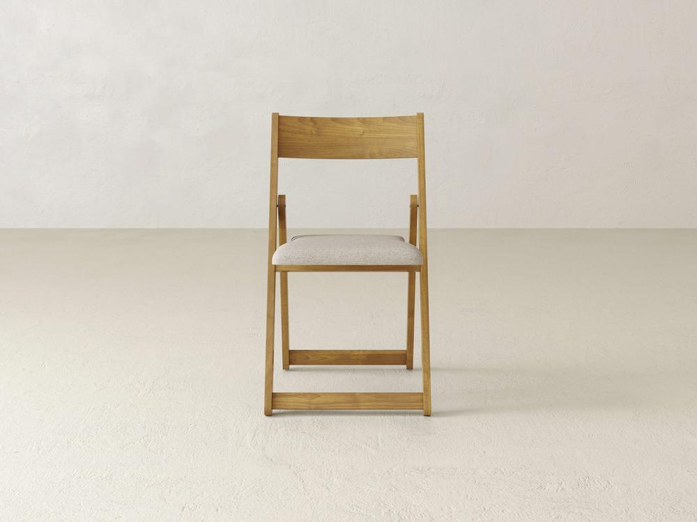 A simple wooden folding chair with a beige cushioned seat is placed on a light-colored floor against a plain, pale wall. The chairs design is minimalist, featuring clean lines and a natural wood finish.