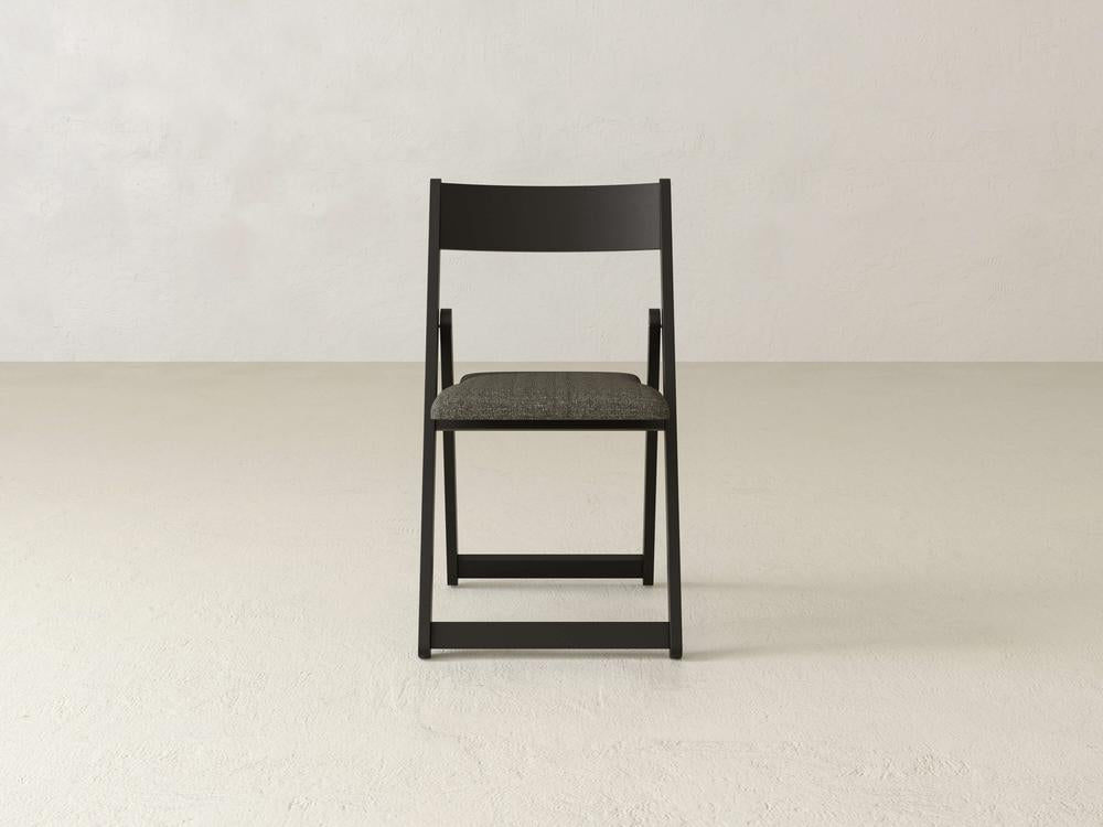 A minimalist black folding chair with a gray fabric seat is placed on a plain, light-colored floor against a bare, off-white wall. The chair is centered and viewed from the front.