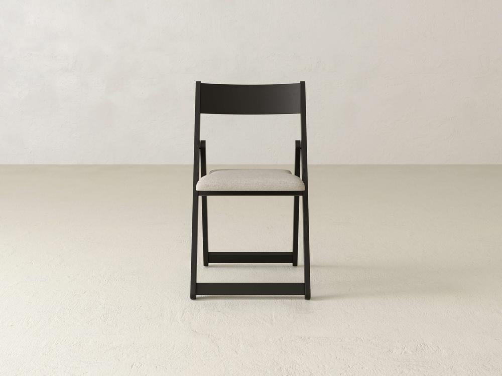 A minimalist black wooden folding chair with a light beige cushioned seat is centered against a plain white textured background. The chair is shown from the front, highlighting its simple and functional design.