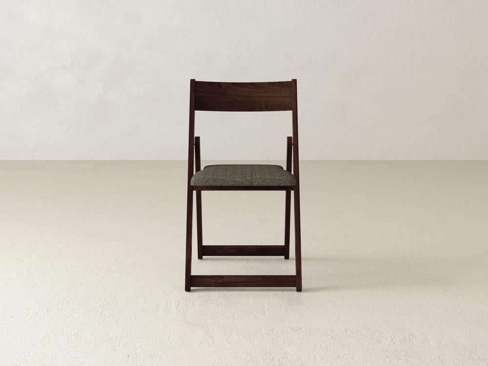 A wooden folding chair with a dark finish and a cushioned gray seat is positioned on a light-colored floor against a plain off-white wall.