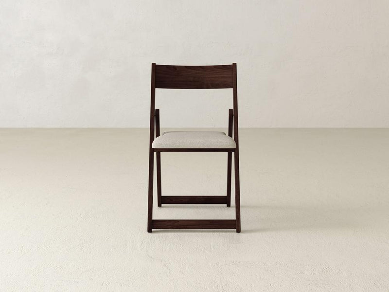 A wooden folding chair with a beige cushioned seat is placed on a light-colored floor against a plain white wall. The chair has a simple and minimalist design.