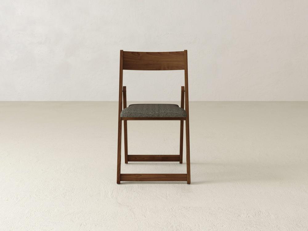 A wooden folding chair with a gray cushioned seat, placed in the center of a minimalist room with white walls and a light-colored floor. The chair is viewed from the front.