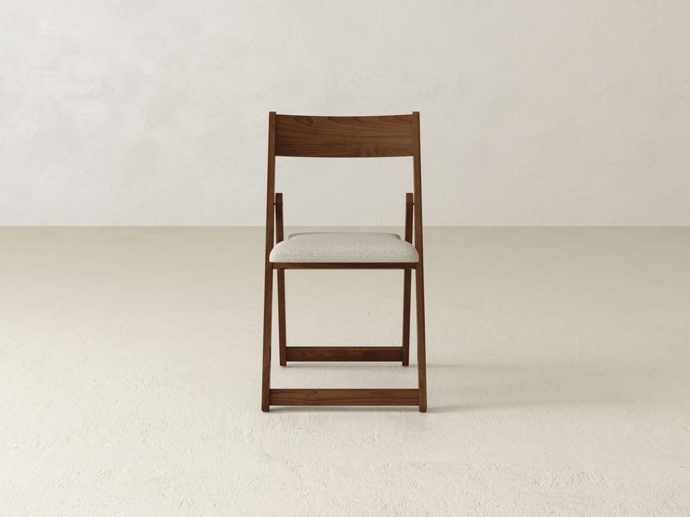 A wooden folding chair with a beige cushioned seat is centered against a plain light backdrop. The simple design features clean lines and a natural wood finish.