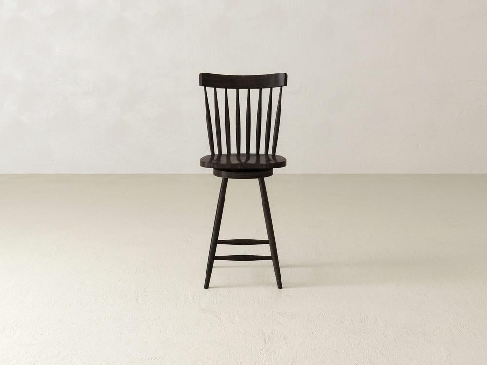 The Rustic Windsor Swivel Stool by James & James, crafted from sturdy maple with an elegant spindle backrest, graces the light beige floor and pale wall. It features a round seat and four splayed legs connected by two footrests.
