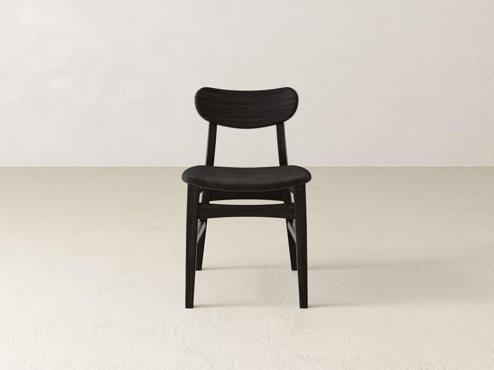 A minimalist, black wooden chair with a curved backrest and simple design sits on a plain, light-colored floor against a white wall.