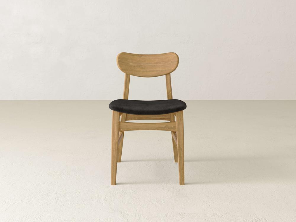 A wooden chair with a black cushioned seat is centered in a minimalist room. The chair has a curved backrest and tapered legs, placed on a light-colored floor against a plain white wall.