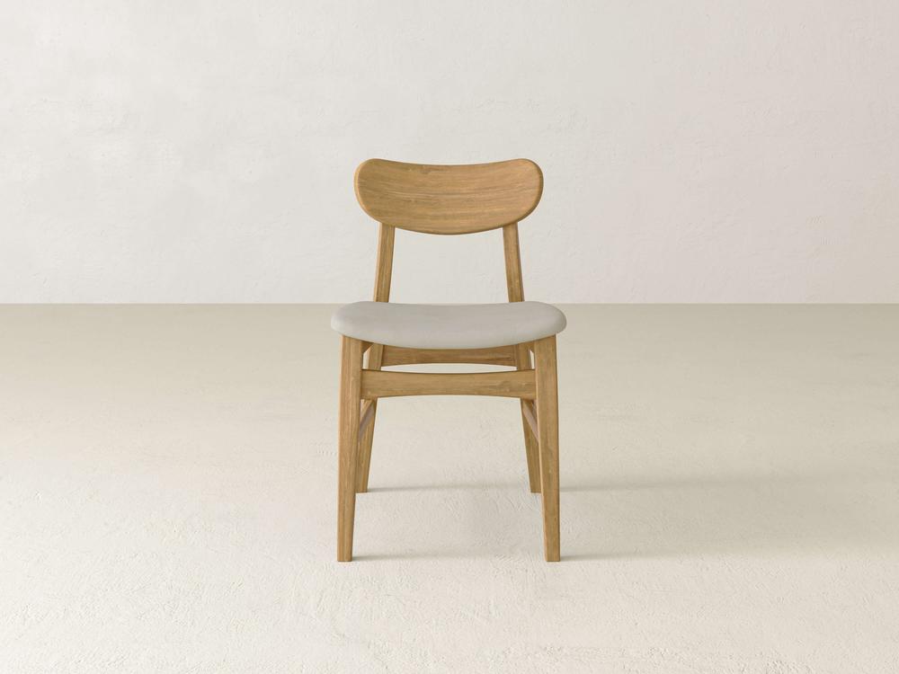 A minimalist wooden chair with a light gray cushioned seat is centered against a plain, light-colored wall and floor background. The chair has a simple, modern design with a slightly curved backrest.