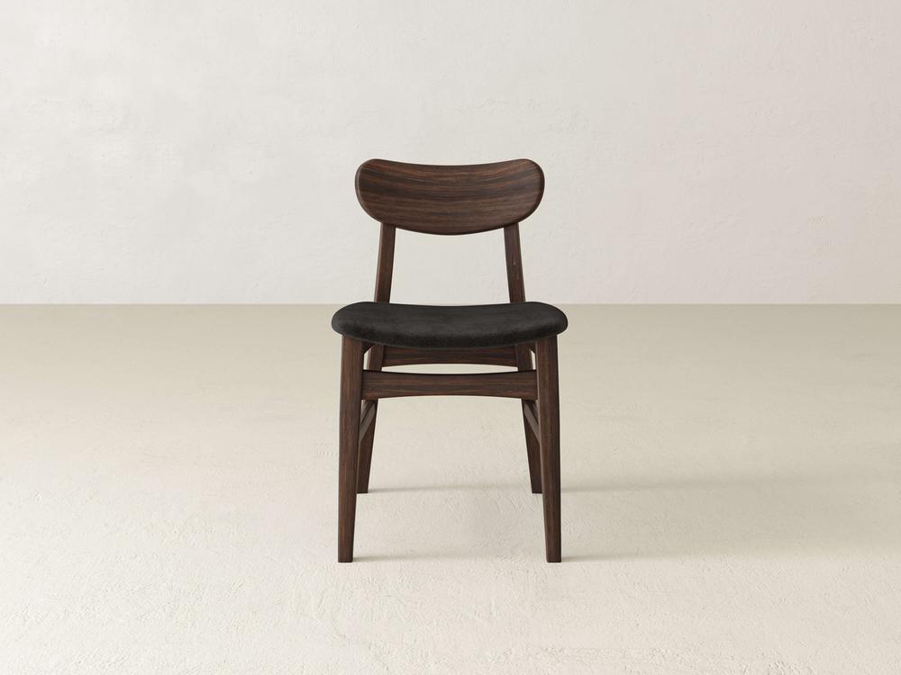 The Lynn Upholstered Dining Chair in Tobacco from James+James, featuring a dark wooden frame and a curved backrest with a black cushioned seat, stands on a plain, light-colored floor against a light-colored wall. The chair boasts a simple, minimalist design with tapered legs.