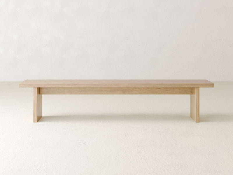 The Noelle Bench by James & James is a minimalist piece crafted from solid white oak, boasting a light natural finish. With its flat top and straight legs, it elegantly complements any modern dining space against a plain, light-colored wall.