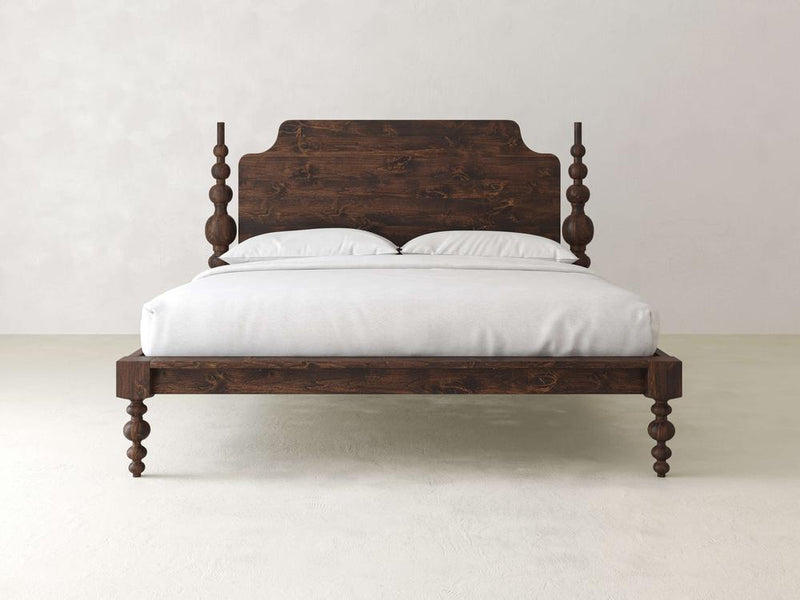 The Olivia Poster Bed from James+James features exquisite craftsmanship with a dark wooden frame and decorative carved posts. It includes an adjustable mattress and white bedding with pillows, all set against a simple light-colored wall and floor.
