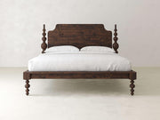 The Olivia Poster Bed from James+James features exquisite craftsmanship with a dark wooden frame and decorative carved posts. It includes an adjustable mattress and white bedding with pillows, all set against a simple light-colored wall and floor.