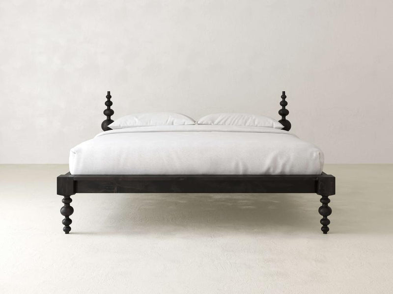 A minimalist bedroom showcases the Olivia Platform Bed - Charred Ember by James+James, with its dark wooden spindle design. It's neatly dressed in white bedding against a plain, light-colored wall and floor for a clean, modern look.