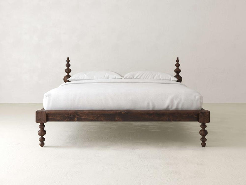 The James+James Olivia Platform Bed, with intricately carved posts, stands against a white wall, featuring neatly arranged white bedding and two pillows for a minimalist and elegant look ideal for an adjustable mattress.