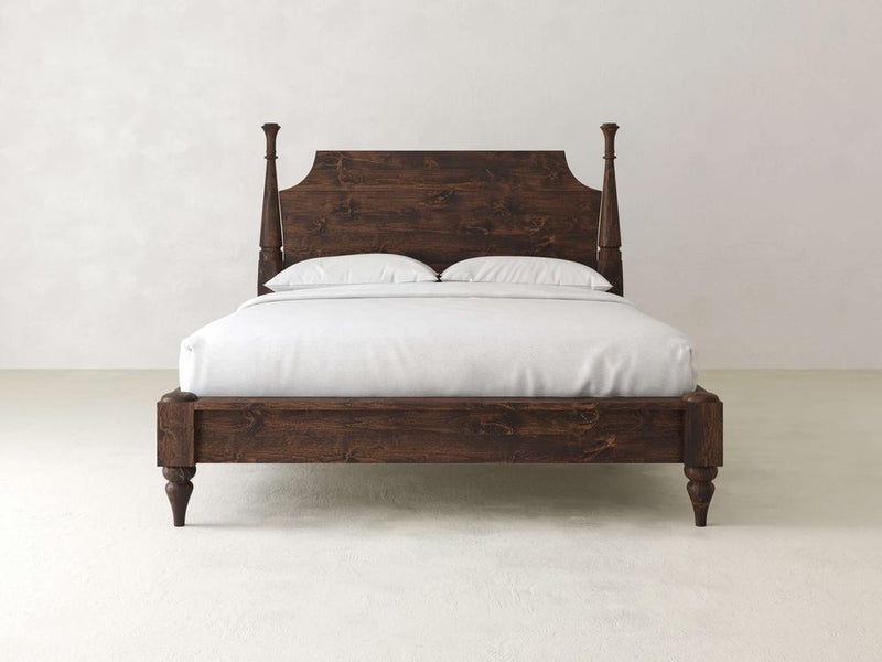 The Stella Bed by James+James features a timeless design with an intricately crafted tall headboard and matching footboard. Adorned with white linens and pillows, it stands elegantly against a plain light-colored wall and floor, highlighting its superior craftsmanship.