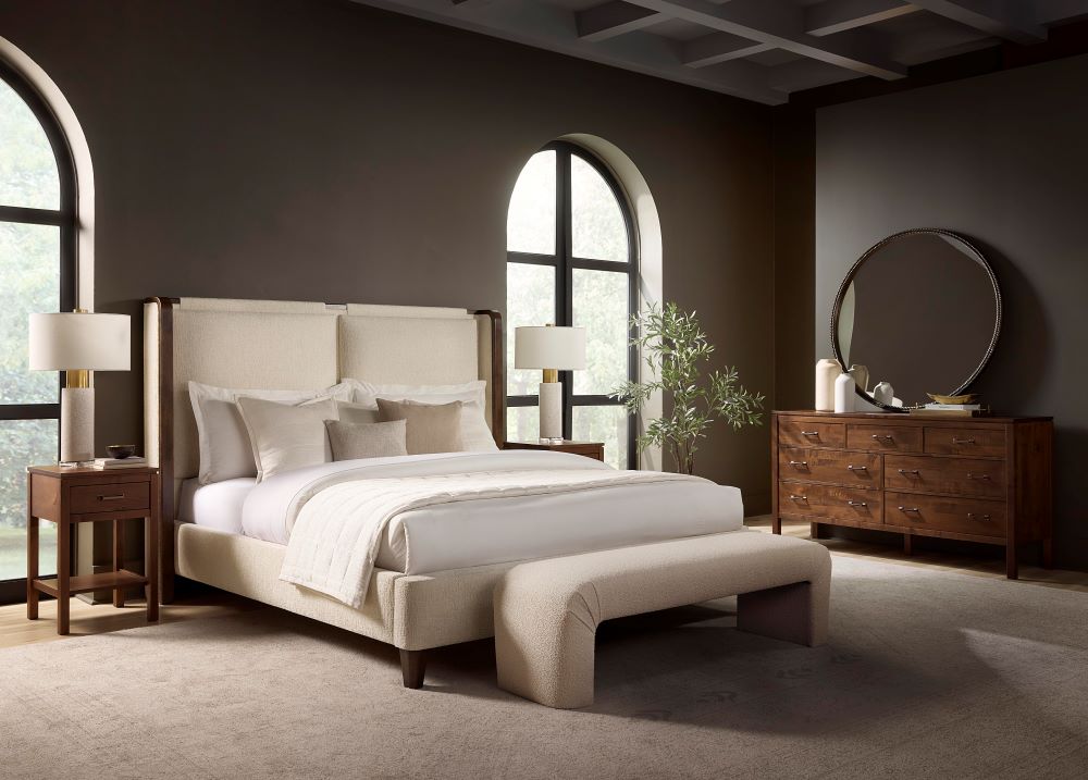 A modern bedroom highlights contemporary design with a James & James Holden Bed in beige leather, dressed in white bedding. Two bedside tables with lamps flank the bed, while a round mirror sits above a maple wood dresser, complemented by large arched windows and a potted plant for natural light and greenery.