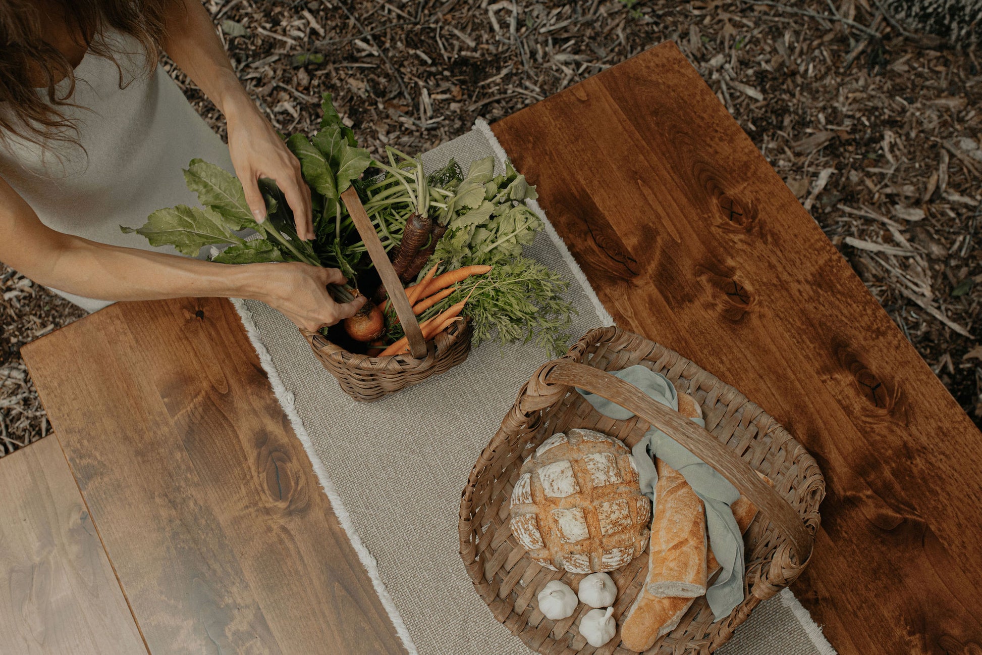 On a Trestle Dining Table - Tobacco by James+James, someone places fresh vegetables, including carrots, in a woven basket. Nearby, another basket holds round and baguette bread plus garlic bulbs. The scene is outdoors.
