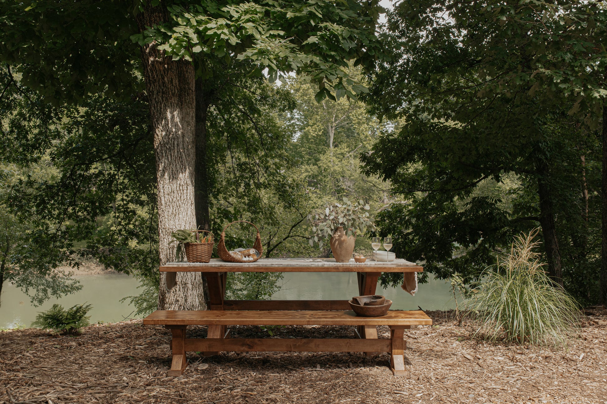 The James+James Trestle Bench in Harvest Wheat on Alder sits under leafy trees by a calm river, set with dishes and baskets, surrounded by greenery and mulched ground.