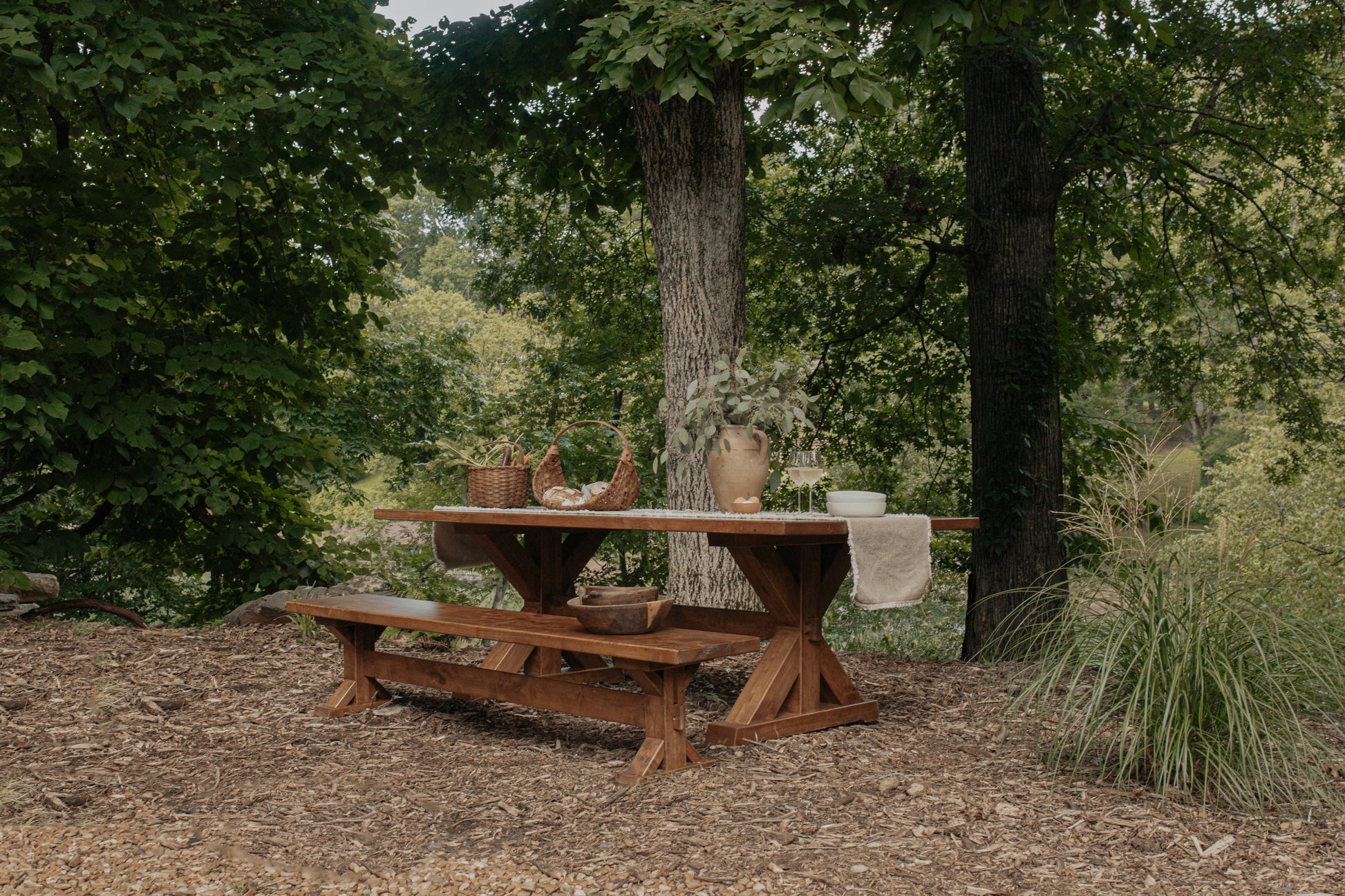 A James+James Trestle Bench in Harvest Wheat on Alder stands outdoors in a wooded area, surrounded by trees and greenery, with baskets, a vase of foliage, and dishes arranged on the rustic wooden table.