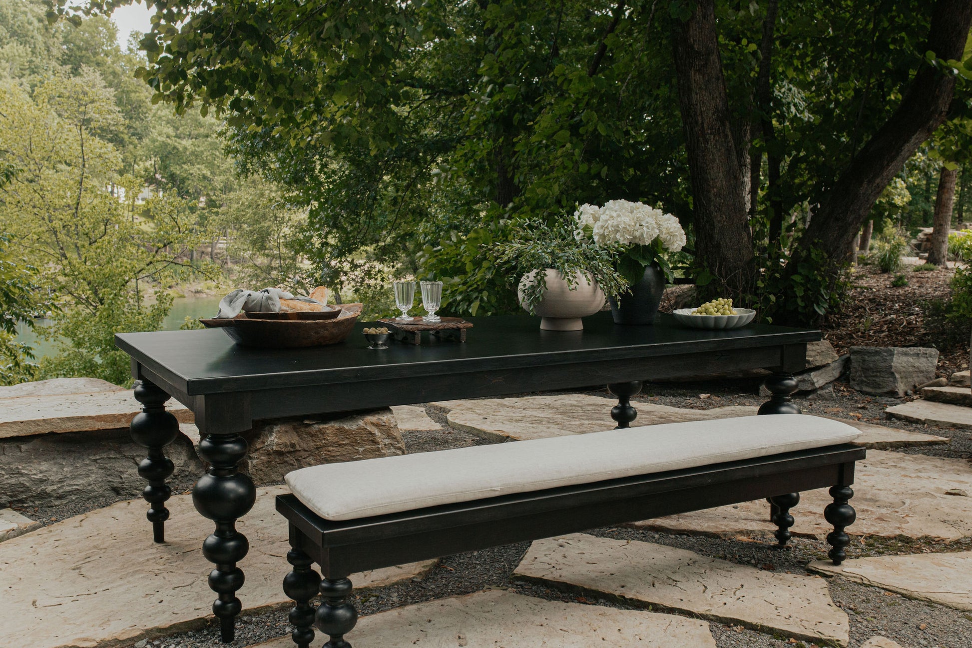 A James+James Olivia Bench in Harvest Wheat on Alder sits outdoors on stone pavers, paired with a black wooden picnic table, both styled with flowers, wine glasses, and snacks among lush green trees and plants.
