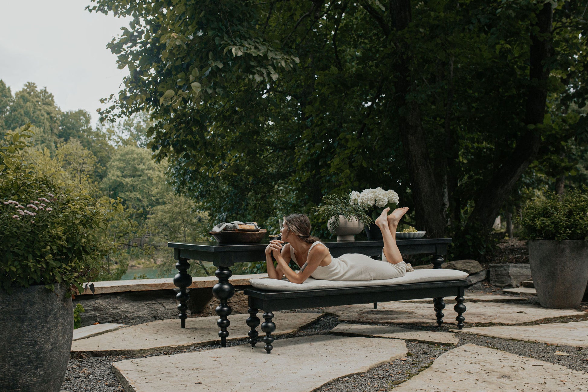 A woman relaxes on the James+James Olivia Bench in Harvest Wheat on Alder, lying on her stomach amid lush garden greenery, holding a mug. Nearby are flowers and food on a table among trees and plants.