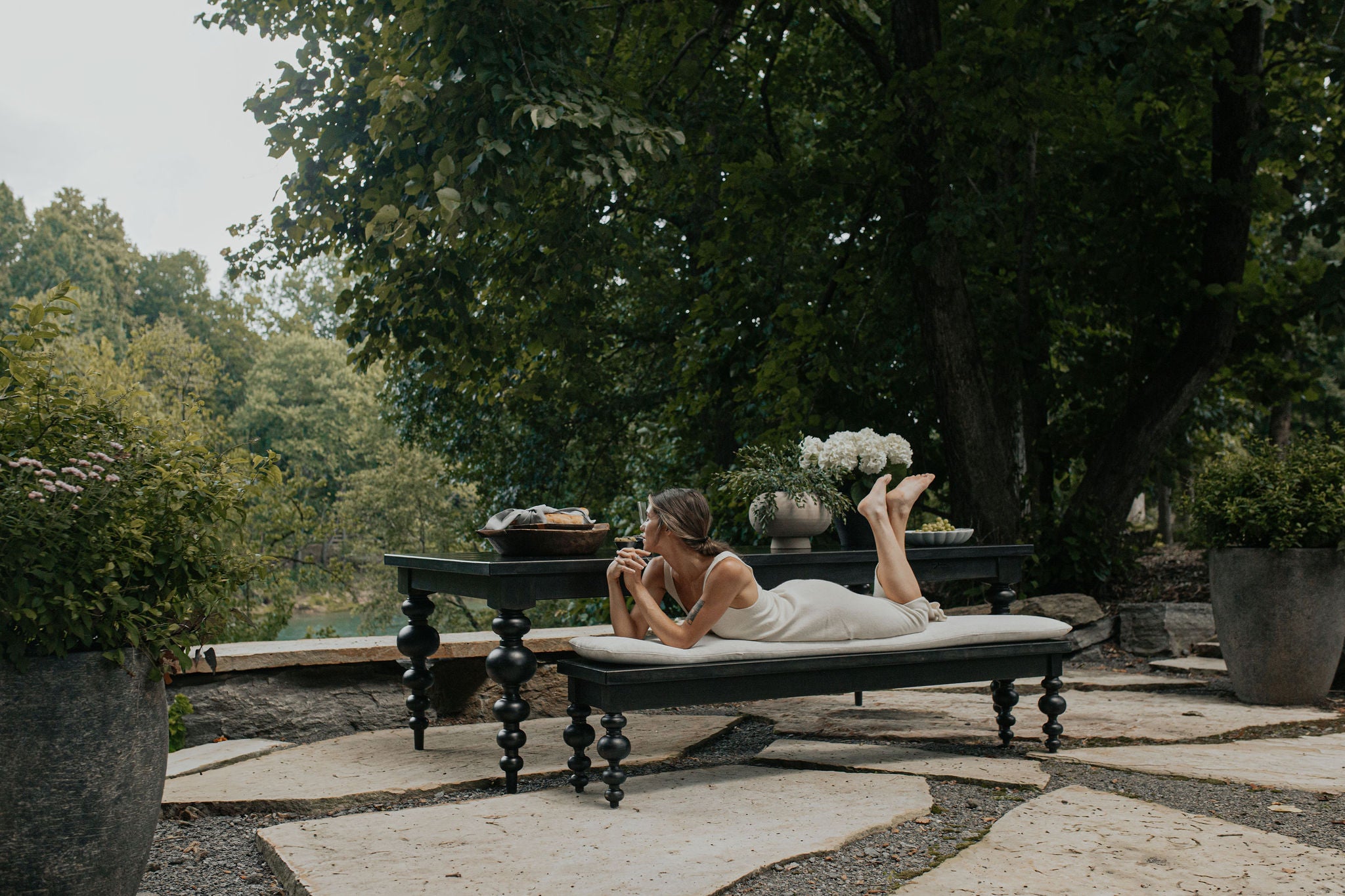 A woman relaxes on the James+James Olivia Bench in Harvest Wheat on Alder, lying on her stomach amid lush garden greenery, holding a mug. Nearby are flowers and food on a table among trees and plants.