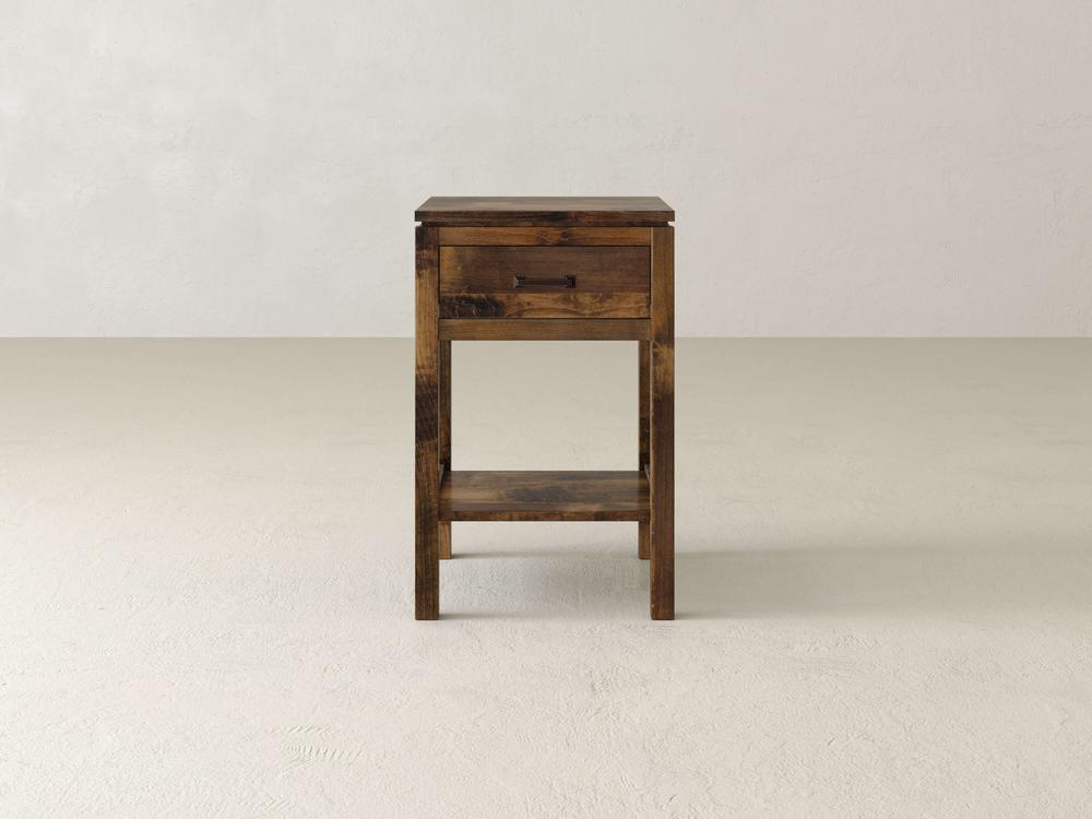 The James & James Jensen 1 Drawer Nightstand, made from solid maple hardwood, epitomizes modern simplicity with a single drawer and open shelf. It leans against a plain, light-colored wall, reflecting simple rustic charm.