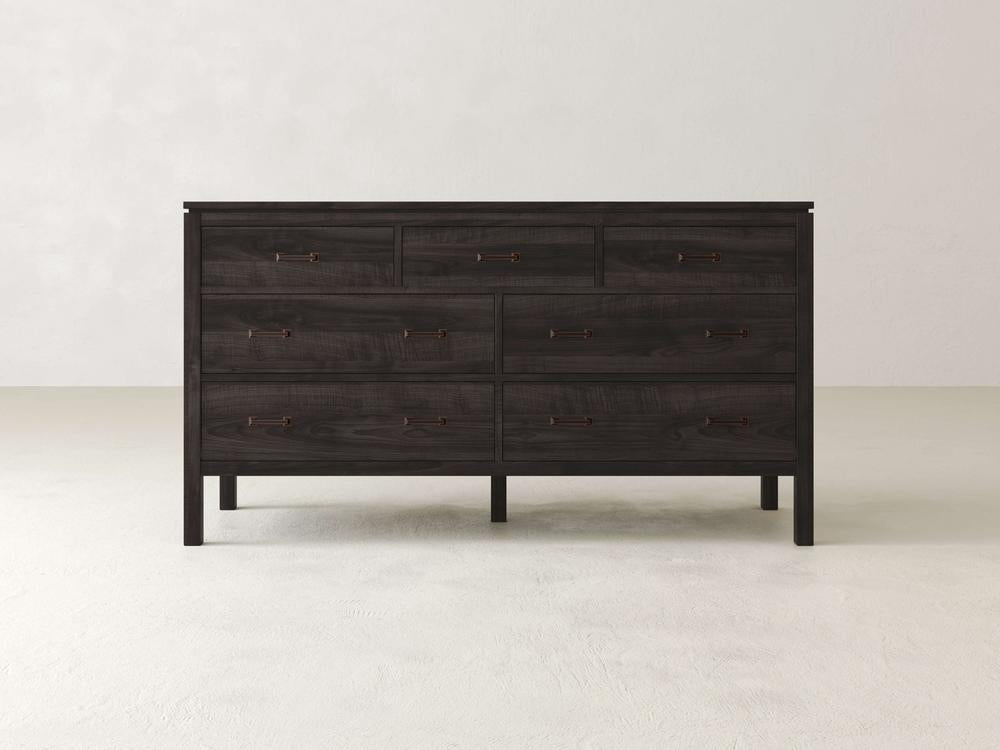 The James & James Jensen 7 Drawer Dresser, crafted from solid maple hardwood with six large drawers and two smaller top ones, features simple metal handles. Its dark wood contrasts against a light wall on a smooth floor, adding a contemporary touch to the space.