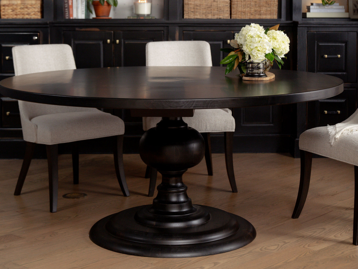 A round dark wooden dining table with a central pedestal base is surrounded by three beige upholstered chairs. The table has an arrangement of white flowers in a small vase. In the background, there is a black cabinet with baskets and a plant.