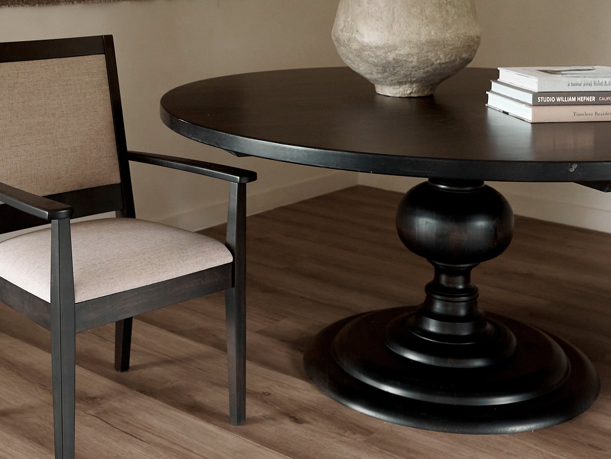 The Josephine Round Dining Table by James+James in Tuscany finish stands next to a matching chair with a beige upholstered seat and back. Books and a stone vase rest on the tabletop, set on medium-toned wood flooring.