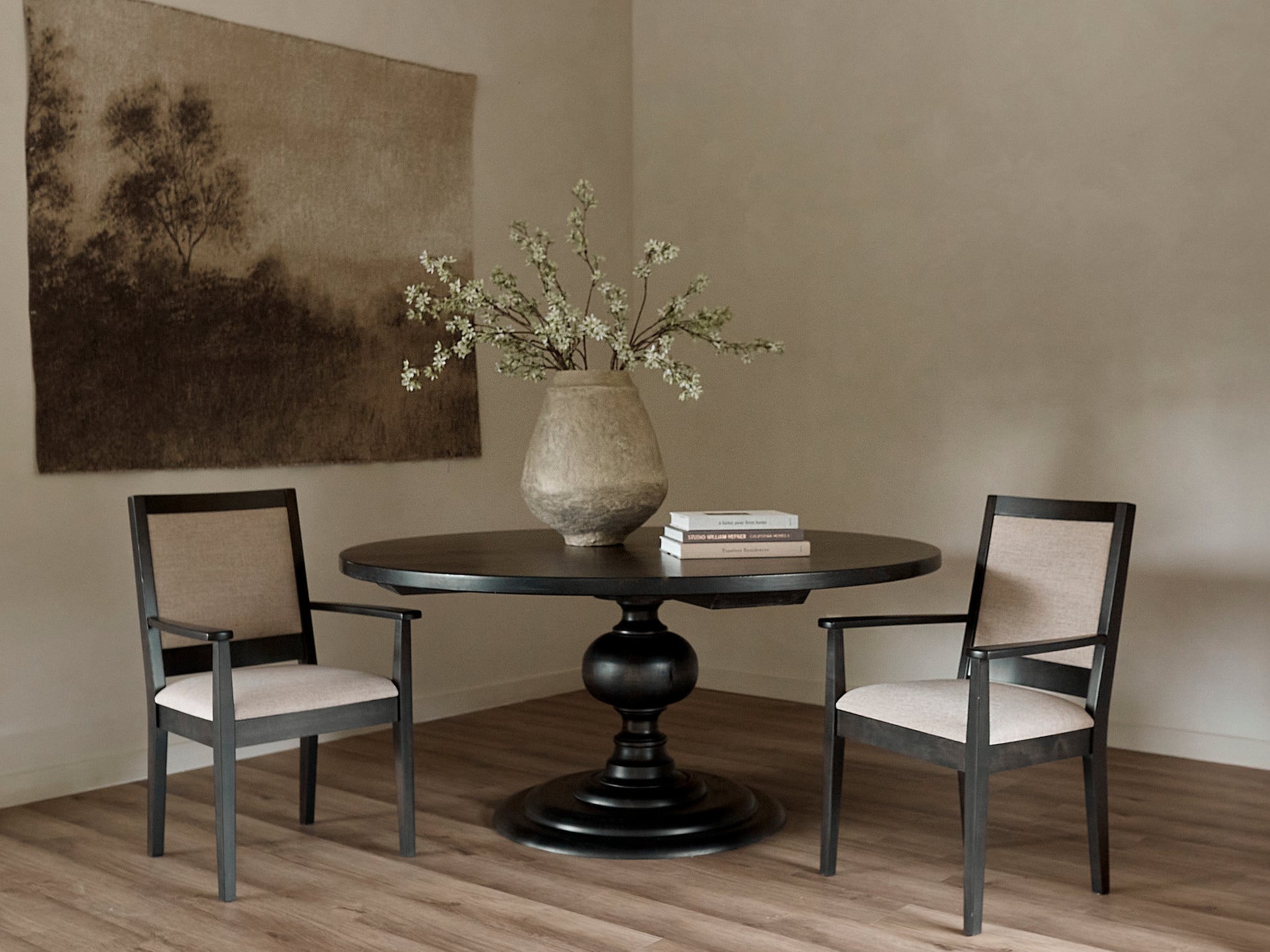The Josephine Round Dining Table - Tuscany by James+James sits between two beige upholstered chairs, topped with a large rustic vase of branches and two books. A sepia landscape artwork hangs above the wood flooring.
