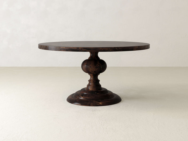 A round, dark wooden pedestal table set against a plain, light-colored background. The table has a thick, ornate central support with a wide, sturdy base.