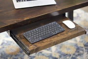 The Standard Desk Add-On by James+James is displayed with a retractable keyboard tray. The tray accommodates a slim, black keyboard and a white wireless mouse. The desk also supports an open laptop, partially visible. The background includes a patterned rug.