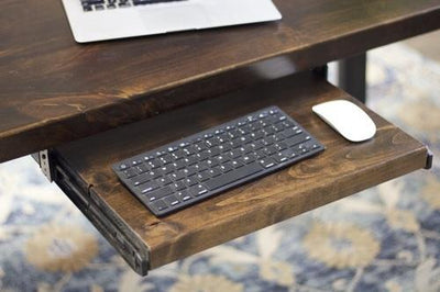 The Standard Desk Add-On by James+James is displayed with a retractable keyboard tray. The tray accommodates a slim, black keyboard and a white wireless mouse. The desk also supports an open laptop, partially visible. The background includes a patterned rug.